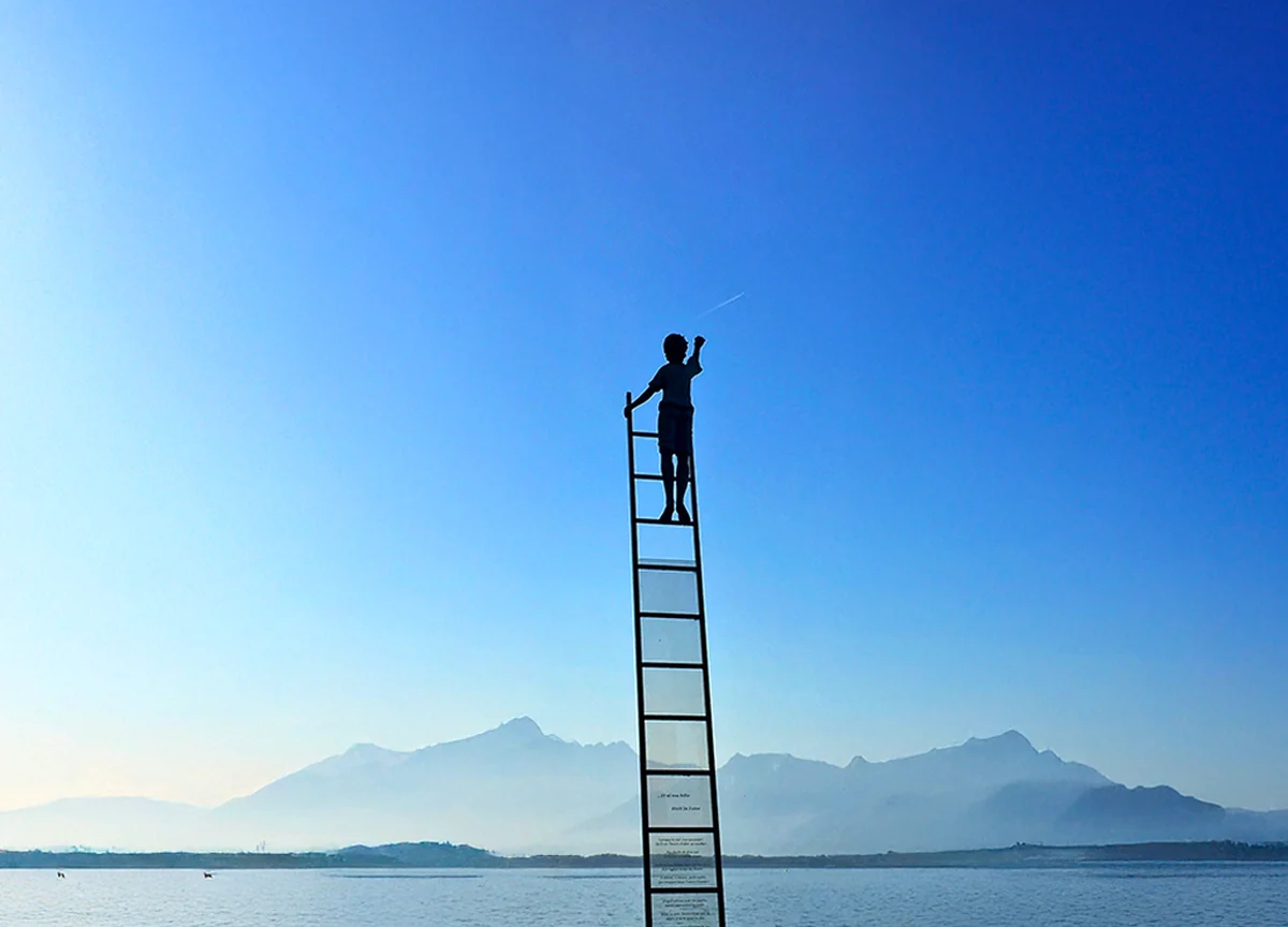 A person at the top of a tall ladder, reaching up against a clear sky